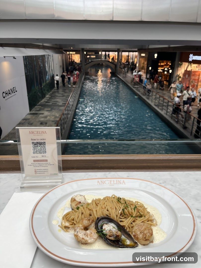A plate of seafood pasta with spaghetti, shrimp, scallops, and mussels in a creamy sauce is presented on a white plate with the Angelina Paris logo. In the background, a canal runs through a shopping mall with various shops and people walking along the waterway. A sign for Angelina with a QR code to order is visible on the table.