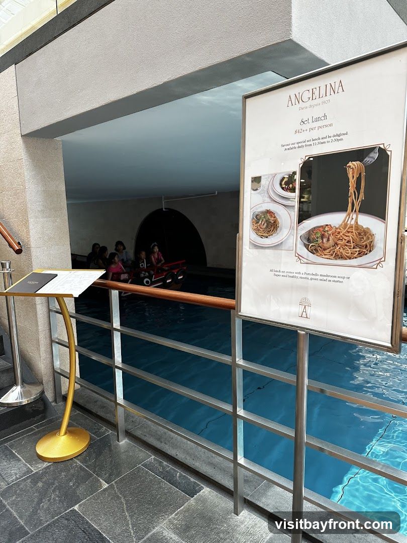 A sign advertising Angelina's set lunch menu is displayed next to a railing overlooking a blue pool of water. The sign features the Angelina logo, the text "Paris depuis 1903", and details about the "Set lunch" priced at "842++ per person". It also includes images of pasta dishes and mentions that the lunch set includes a Portobello mushroom soup or a super seed healthy ricotta green salad as a starter. The sign indicates the lunch is available daily from 11:30 am to 2:30 pm.