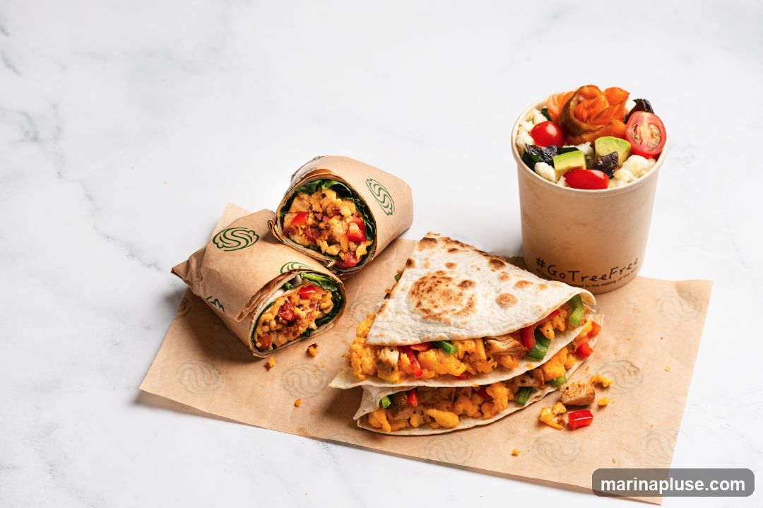 Two wraps, one cut in half, and a folded quesadilla are arranged on brown paper. A cup filled with salad and topped with a carved carrot rose is to the right. The paper and cup have a green logo and the hashtag #GoTreeFree.