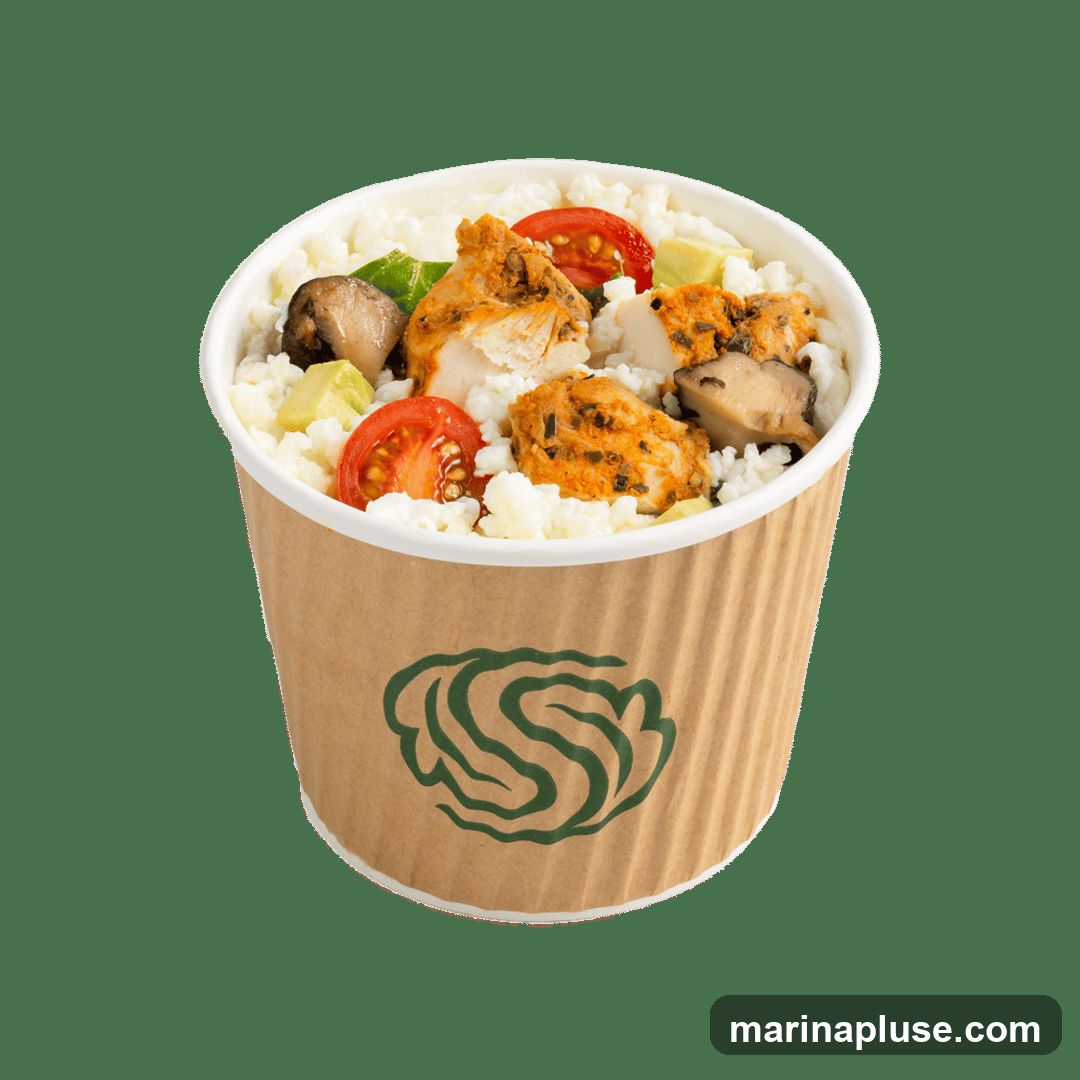 A close-up studio shot of a take-out bowl filled with white rice, grilled chicken pieces, cherry tomato halves, avocado cubes, and mushrooms. The bowl is made of corrugated brown paper with a green logo printed on the side. The background is black.