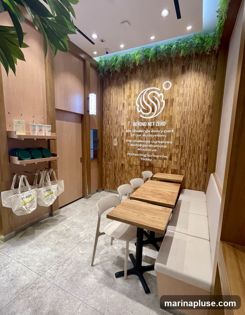 The image shows the interior of a cafe or restaurant with a seating area. The walls are decorated with wooden panels and greenery. A large logo and text that reads "BEYOND NET ZERO" are displayed on the back wall. There are several tables and chairs, as well as a banquette seat. On the left, there are shelves with cups and tote bags.