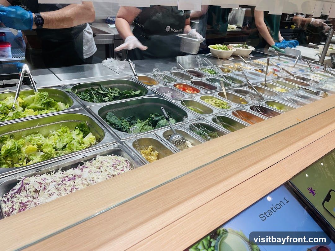 A salad bar with various fresh ingredients displayed in metal containers. Options include lettuce, spinach, kale, shredded cabbage, tomatoes, edamame, and pasta. Staff members in aprons and gloves are visible in the background, preparing food. A digital screen labeled "Station 1" is in the foreground.