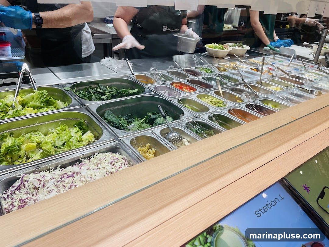 A salad bar with various fresh ingredients displayed in metal containers. Options include lettuce, spinach, kale, shredded cabbage, tomatoes, edamame, and pasta. Staff members in aprons and gloves are visible in the background, preparing food. A digital screen labeled "Station 1" is in the foreground.