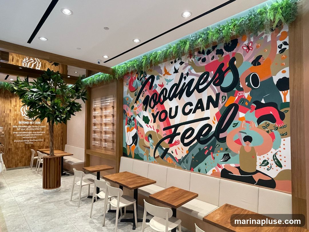 The interior of a cafe features a large, colorful mural on the wall with the text "Goodness YOU CAN Feel". The mural depicts abstract figures and nature elements. The cafe has wooden tables and chairs, with banquette seating along the mural wall. A large artificial tree stands near the entrance. A wooden panel on the left wall has text about "BEYOND NET ZERO".