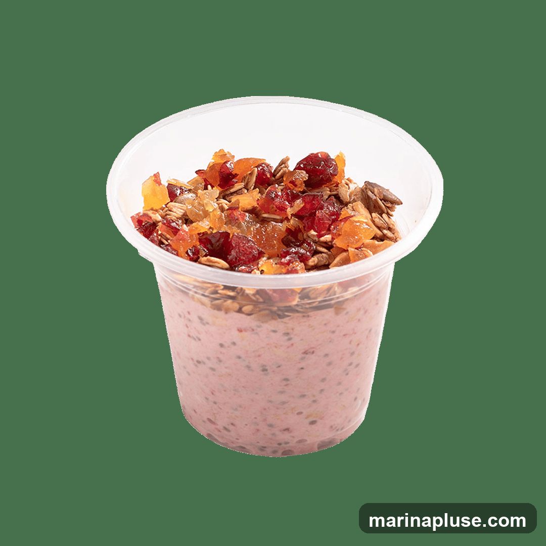 A clear plastic cup filled with pink overnight oats, topped with granola, dried cranberries, and diced dried apricots.