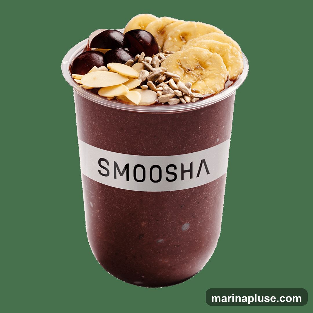 A smoothie bowl in a clear plastic cup with a white band labeled "SMOOSHA". The bowl is topped with sliced bananas, dark berries, sliced almonds, and sunflower seeds.