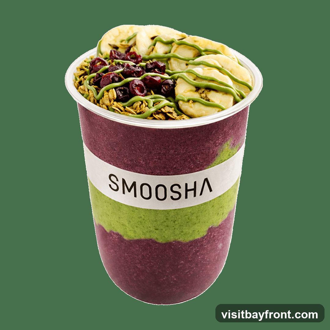 A smoothie bowl in a clear plastic cup with a white band labeled "SMOOSHA". The smoothie is layered with a dark purple base and a green layer. It is topped with granola, dried cranberries, sliced bananas, and a drizzle of green sauce.