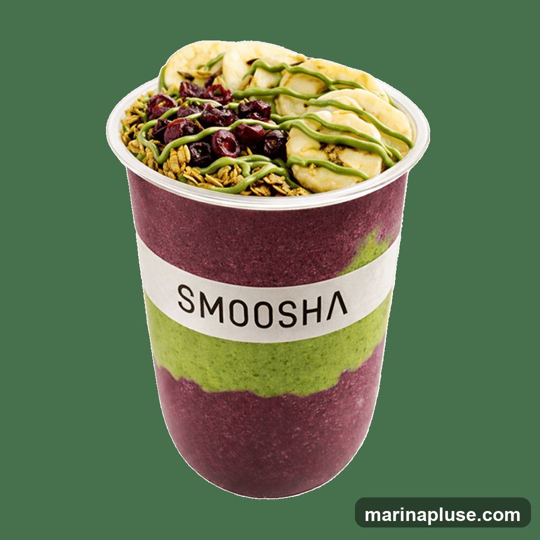 A smoothie bowl in a clear plastic cup with a white band labeled "SMOOSHA". The smoothie is layered with a dark purple base and a green layer. It is topped with granola, dried cranberries, sliced bananas, and a drizzle of green sauce.