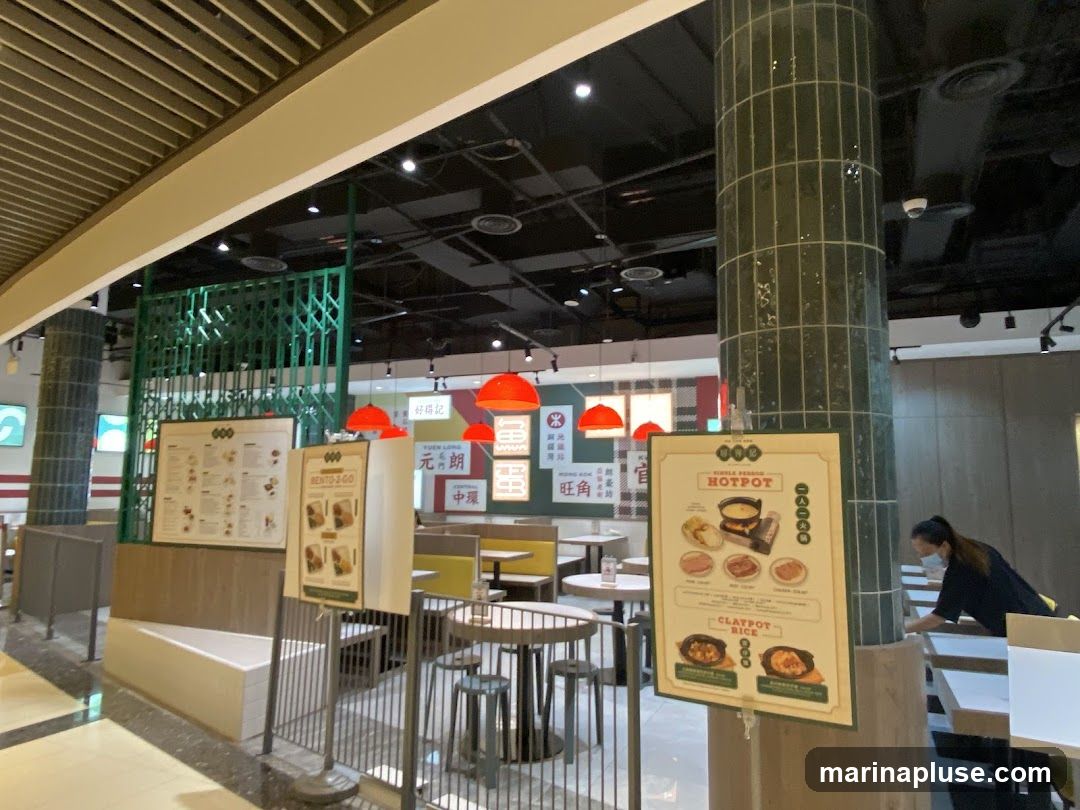 The image shows the interior of a restaurant with several menu boards displayed. One prominent menu board features "HOTPOT" and "CLAYPOT RICE" with images of the dishes. Another board displays "BENTO-2-GO". Red pendant lights hang from the ceiling. Tables and seating are visible, along with a person in the background wearing a mask.