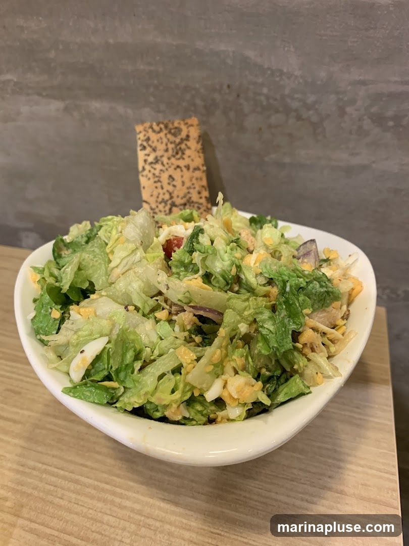 A close-up shot of a salad in a white bowl, topped with shredded cheese, chopped eggs, and red onion. A cracker with poppy seeds is stuck into the salad. The background is a textured gray wall, and the bowl sits on a wooden surface.