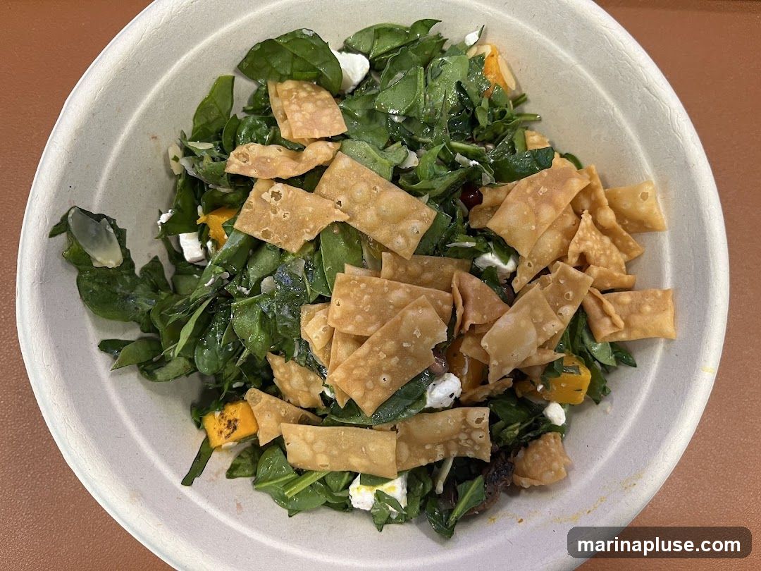 A close-up overhead shot of a salad in a compostable bowl. The salad contains leafy greens, cubes of white cheese, roasted squash, and crispy fried wonton strips.
