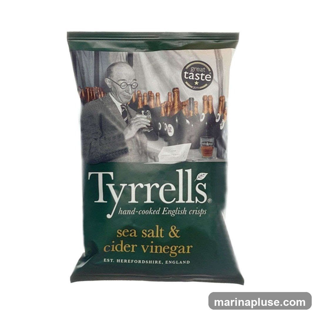 A bag of Tyrrells Sea Salt & Cider Vinegar hand-cooked English crisps. The packaging features a black and white illustration of a man in glasses holding a drink, with bottles in the background. It also has a "Great Taste 2019" award logo. The flavor is prominently displayed as "sea salt & cider vinegar" and the origin as "EST. HEREFORDSHIRE, ENGLAND".