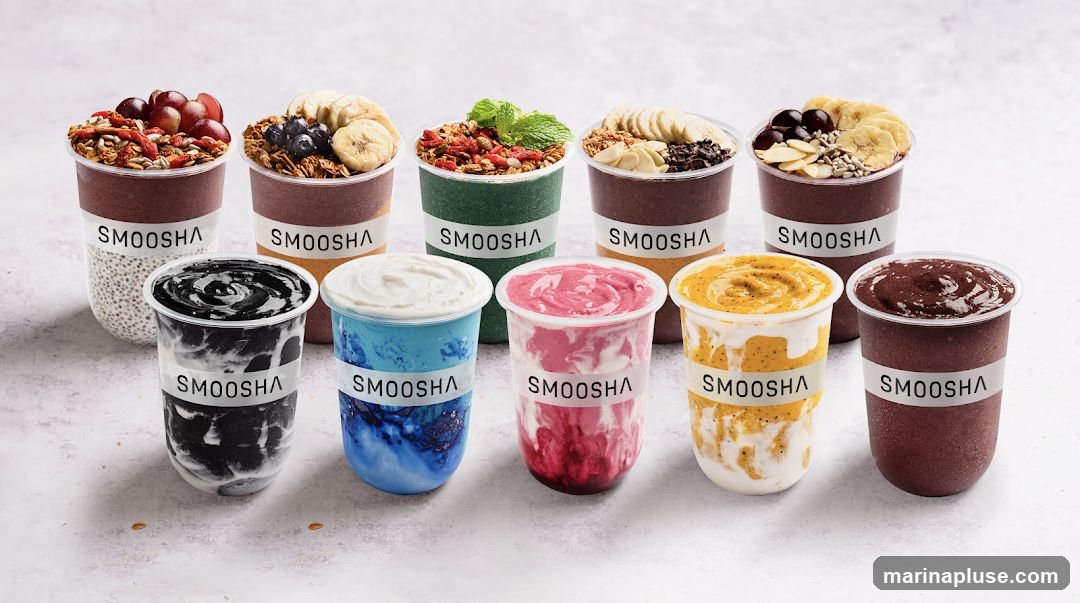 A collection of eight smoothie bowls and drinks in clear plastic cups, each with a white band labeled "SMOOSHA". The bowls at the back are topped with granola, fruits like grapes, blueberries, and bananas, and seeds. The drinks in the front are various colors, including black, blue, pink, yellow, and dark purple, with swirled textures.