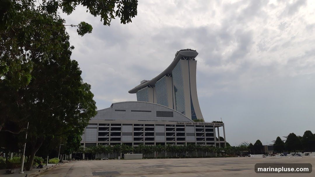 A wide shot of the Marina Bay Sands Expo and Convention Centre building under a cloudy sky. The iconic hotel structure with its boat-shaped top is visible behind the convention center. Trees line the left side of the frame, and a large, empty paved area is in the foreground.