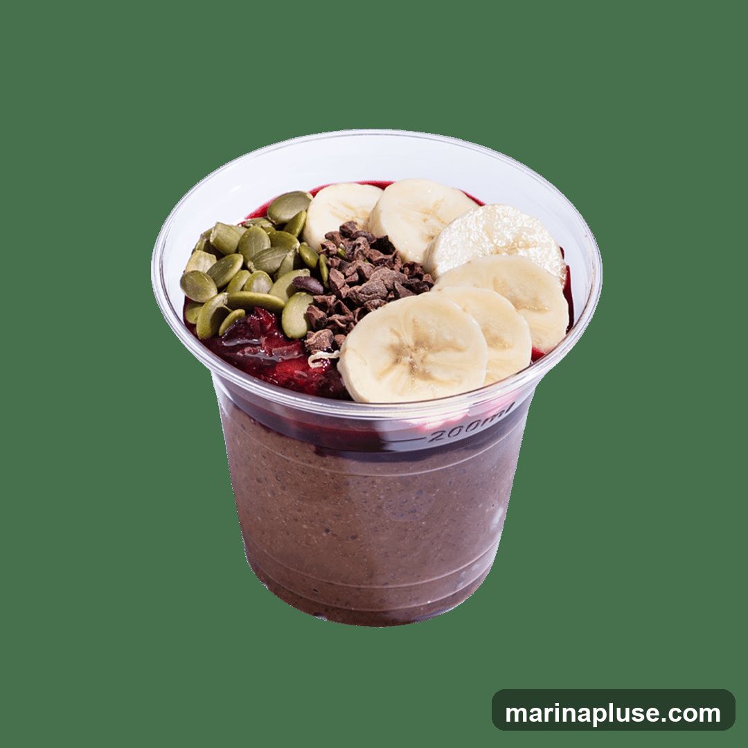 A chocolate smoothie bowl in a clear plastic cup, topped with sliced bananas, pumpkin seeds, and chocolate chips. The cup has a 200ml marking.