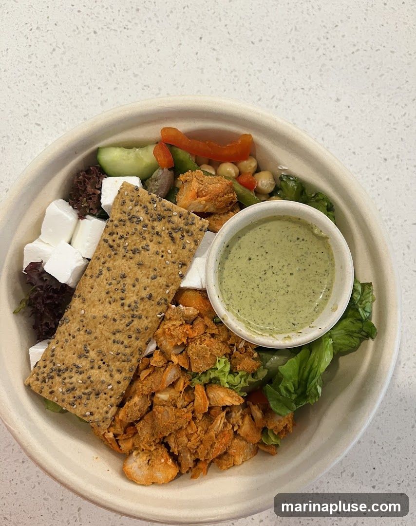 A close-up overhead view of a chicken salad bowl. The bowl contains a generous portion of shredded chicken coated in a reddish-orange spice blend, cubes of white cheese, a large cracker topped with seeds, lettuce, cucumber slices, red bell pepper strips, chickpeas, and a small cup of green dressing. The bowl is made of a light-colored, textured material.