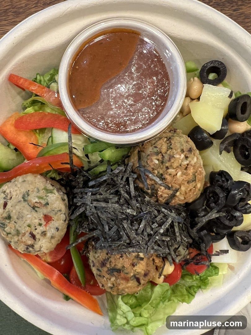 A close-up overhead view of a salad bowl filled with various ingredients. The bowl contains lettuce, cherry tomatoes, sliced red and green bell peppers, black olives, chickpeas, and what appear to be falafel balls. A small white bowl in the center holds two types of dressing, one darker and thicker, the other lighter and more liquid. The falafel balls are topped with shredded seaweed. The bowl is made of a light-colored, textured material.
