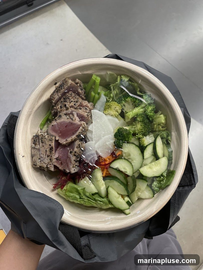 A close-up overhead view of a salad bowl filled with various ingredients. The bowl contains slices of seared tuna, lettuce, cucumber slices, shredded carrots, broccoli florets, and thinly sliced white radish. The salad is covered with a clear plastic lid. The bowl is held within a gray fabric bag.