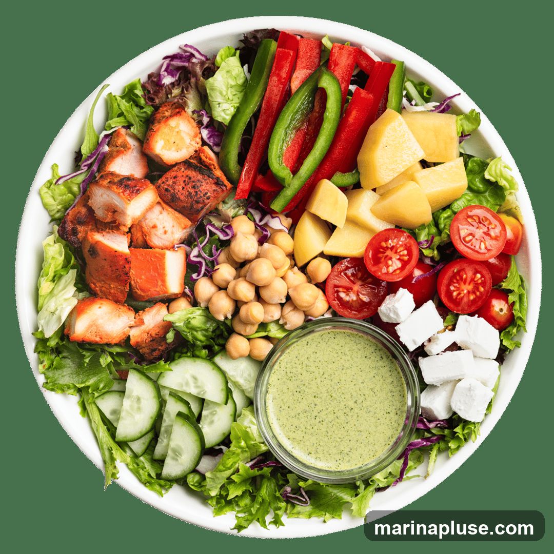 A white bowl filled with a colorful salad. The salad contains grilled chicken pieces, chickpeas, cucumber slices, cherry tomatoes, feta cheese cubes, diced pineapple, and sliced red and green bell peppers, all on a bed of lettuce and red cabbage. A small glass bowl of green dressing is in the center.