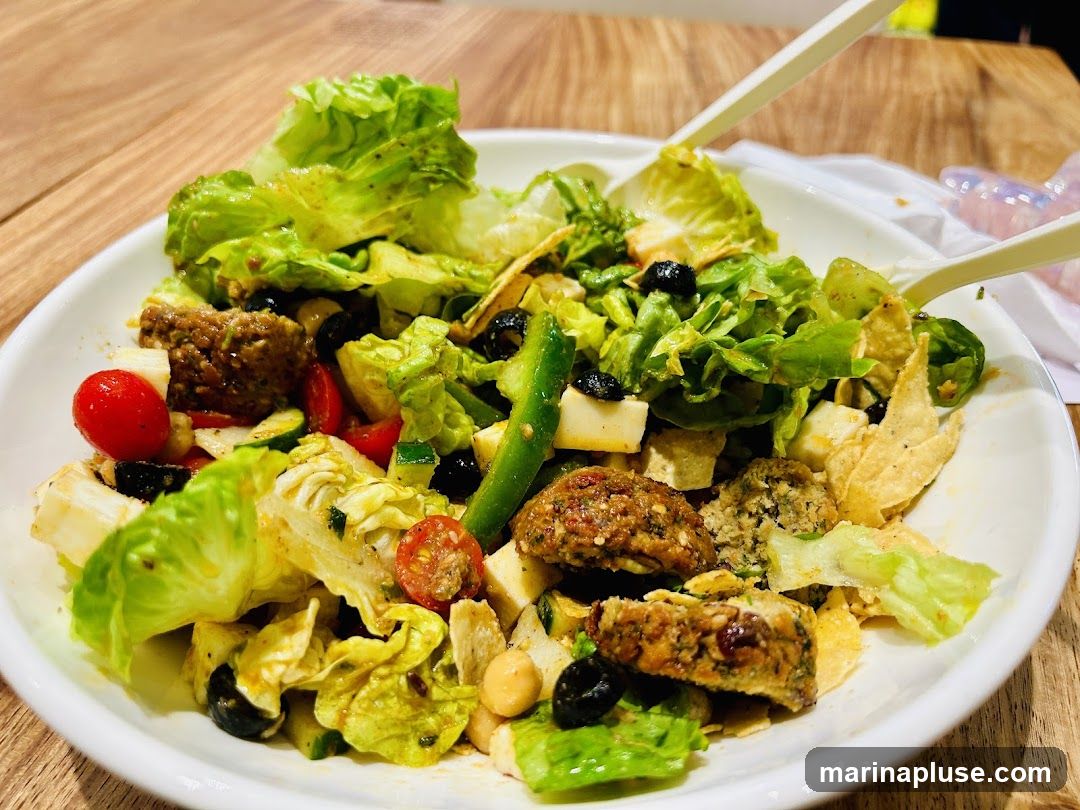 A close-up shot of a white bowl filled with a vibrant salad. The salad contains lettuce, cherry tomatoes, olives, cucumber, falafel balls, and cubes of cheese or tofu. Two white utensils are visible, resting on the edge of the bowl. The background shows a wooden surface and a blurred pink object.
