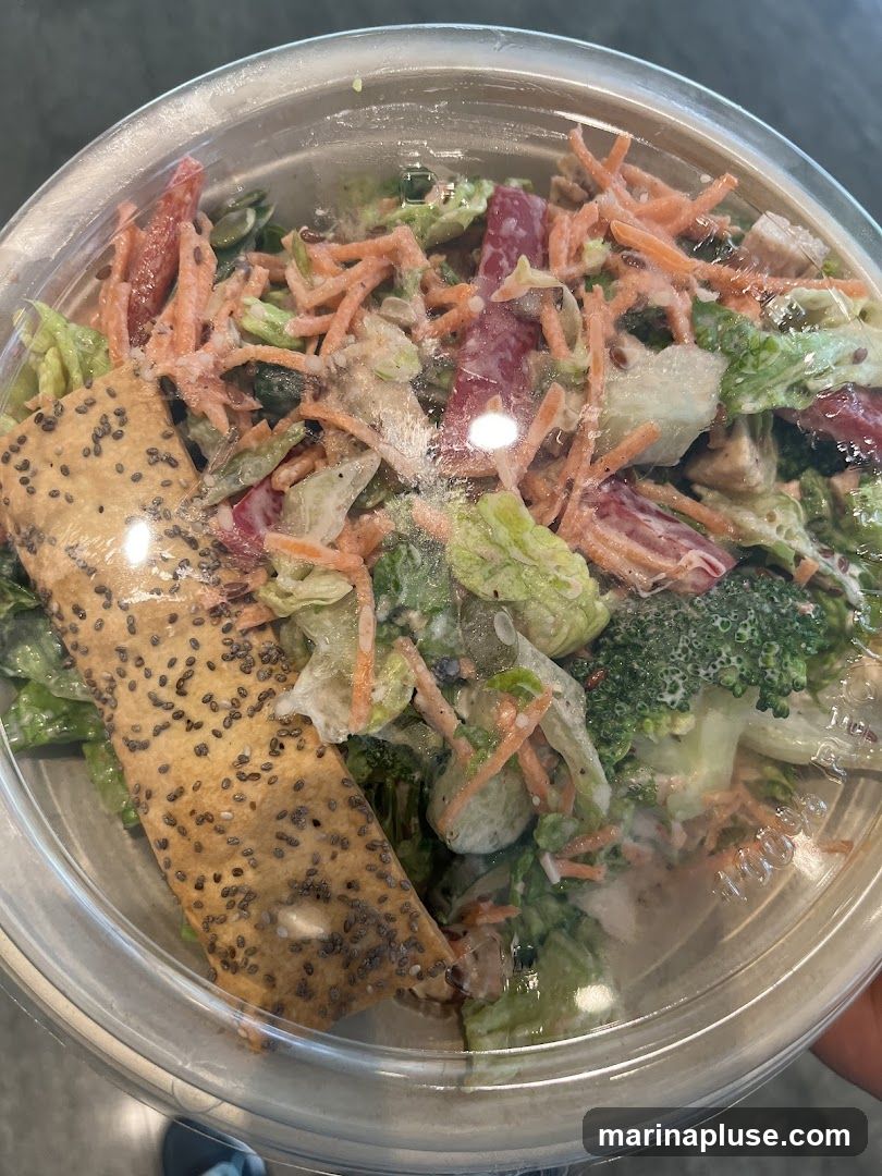 A close-up shot of a salad in a clear plastic container. The salad appears to be made with lettuce, shredded carrots, broccoli florets, and possibly some red peppers. A large cracker or flatbread, sprinkled with chia seeds, is placed on top of the salad. The dressing is creamy and coats the ingredients.