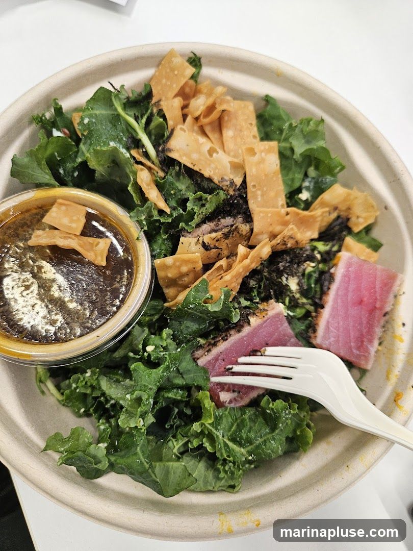 A close-up shot of a tuna salad bowl. The bowl contains a generous portion of kale, seared tuna slices, crispy wonton strips, and what appears to be seaweed. A small container of dressing is also visible. A white plastic fork is placed in the salad.