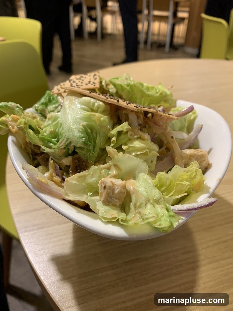 A close-up shot of a chicken salad in a white bowl, placed on a wooden table. The salad contains lettuce, chicken pieces, red onion slices, and is dressed with a creamy sauce. Two crackers topped with chia seeds are placed on top of the salad. In the background, blurred figures of people and chairs are visible, suggesting a dining environment.