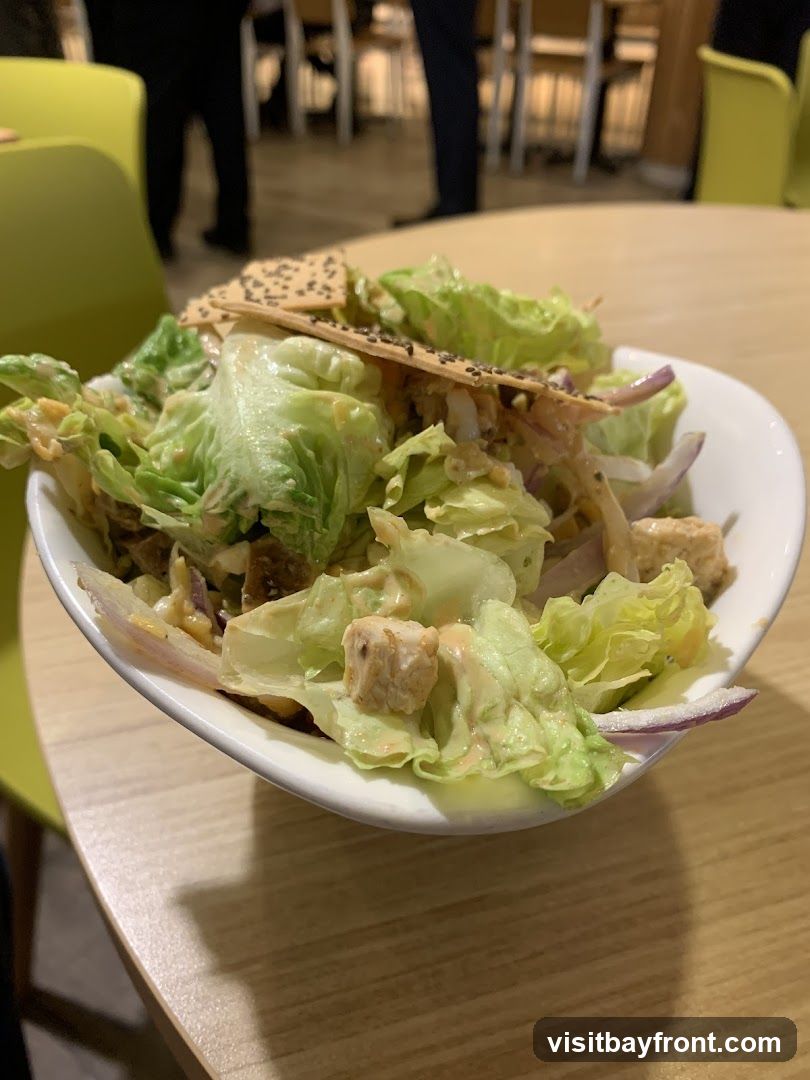 A close-up shot of a chicken salad in a white bowl, placed on a wooden table. The salad contains lettuce, chicken pieces, red onion slices, and is dressed with a creamy sauce. Two crackers topped with chia seeds are placed on top of the salad. In the background, blurred figures of people and chairs are visible, suggesting a dining environment.