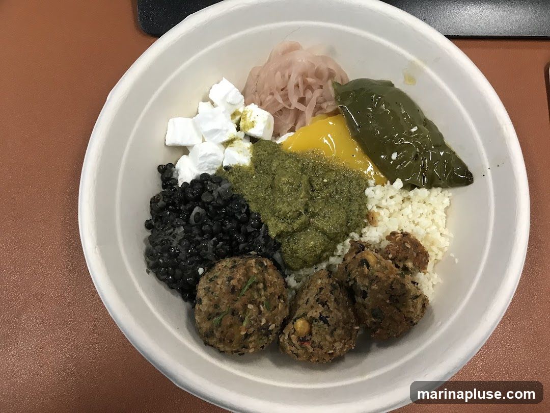 A white bowl filled with various ingredients, including black beans, cubed feta cheese, a green sauce, cauliflower rice, falafel balls, pickled onions, a yellow sauce, and a roasted green pepper.