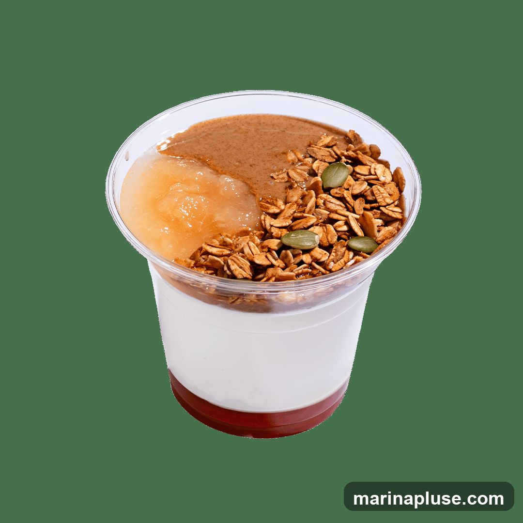 A clear plastic cup filled with layers of yogurt, fruit compote, nut butter, and granola with pumpkin seeds.