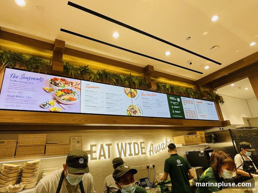 A digital menu board displays various food options, including "The Seasonals" with "Truffle Wonder" and "Amalfi Breeze," and "The Signatures" with items like "Matl Caesar." There are also sections for "Warm Grain Bowls" and "Snacks." Below the menu, staff members in uniform are visible working behind a counter. The restaurant's slogan "EAT WIDE Awake" is illuminated on the wall.
