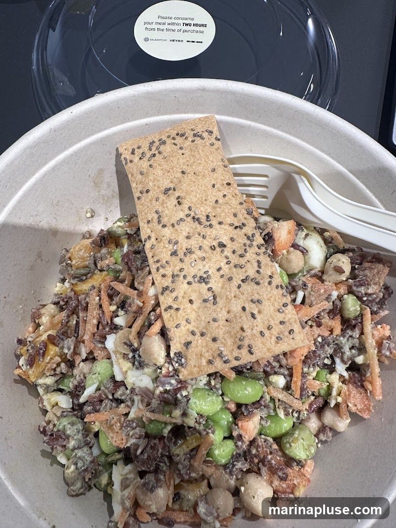 A close-up view of a salad in a compostable bowl, topped with a cracker. The salad appears to contain edamame, shredded carrots, chickpeas, and grains. A clear plastic lid is visible in the background with a circular sticker that reads "Please consume your meal within TWO HOURS from the time of purchase" and logos for "SALADSTOM", "HEYBO", and "UK-MEALS".