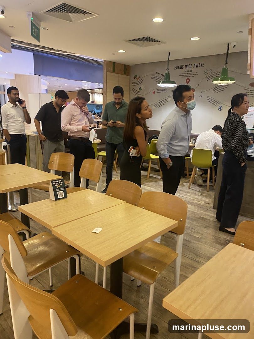 A group of people are standing and waiting in a restaurant. Some are looking at their phones, while others are looking around. The tables and chairs are made of wood and metal. There is a sign on one of the tables with a QR code and the text "TELL US WHAT YOU REALLY THINK". A mural with the text "EATING WIDE AWAKE" is visible on the wall.