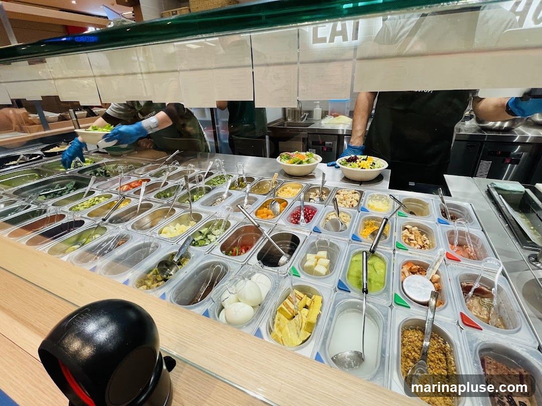 A salad bar with a wide variety of fresh ingredients displayed in individual containers. Staff members in the background are preparing salads. The counter is made of wood, and there is a black scanner in the foreground.