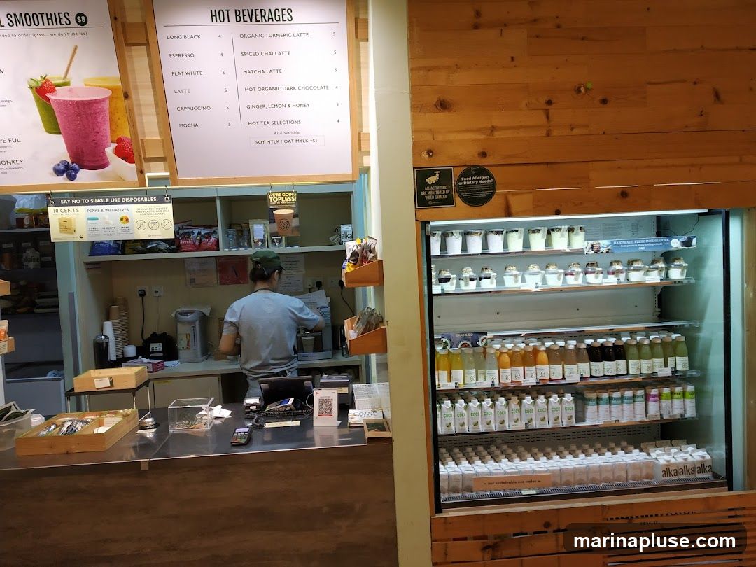 A view of a cafe counter with a person working behind it, and a large refrigerator filled with drinks and desserts. A menu board is visible above the counter, listing hot beverages. Signs about single-use disposables and food allergies are also present.
