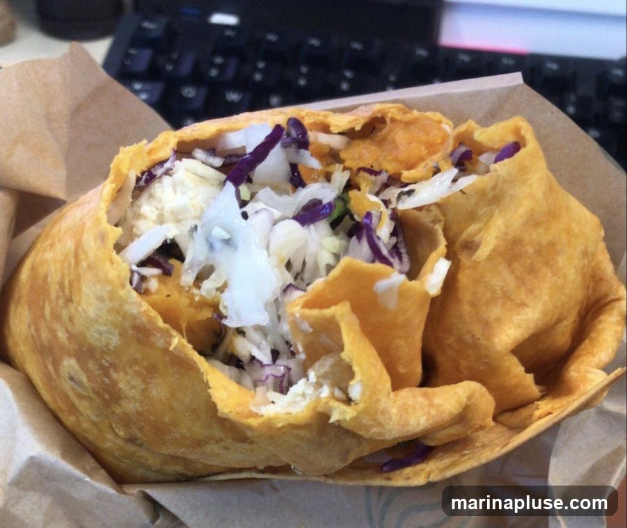 A close-up shot of a golden-brown, flaky pastry filled with a mixture of shredded cabbage, purple cabbage, and white cheese or tofu. The pastry appears to be a type of empanada or turnover, served in a brown paper wrapper. The background shows a blurred keyboard and desk area.