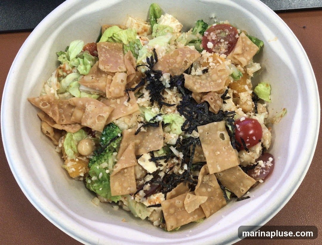 A close-up overhead view of a salad in a white bowl. The salad contains lettuce, cherry tomatoes, broccoli florets, chickpeas, and crispy fried wonton strips. It is topped with shredded nori seaweed and possibly some cheese or tofu cubes. The salad appears to be a healthy and colorful meal.