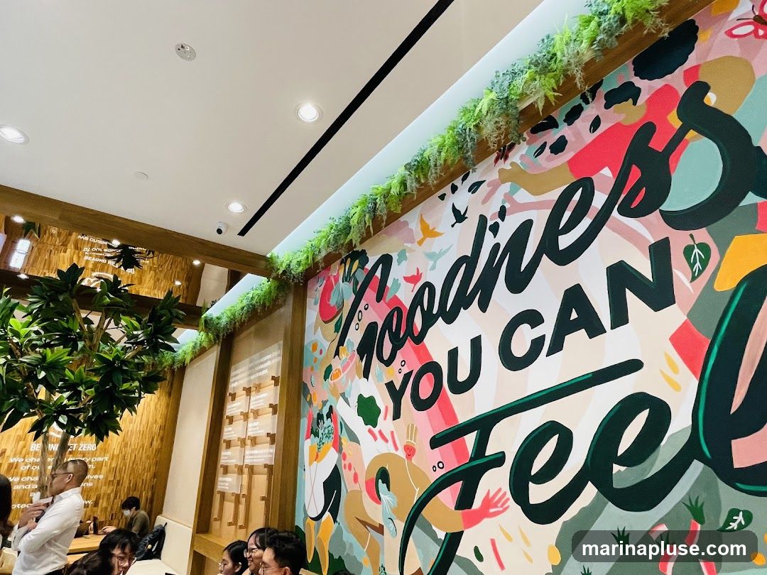A colorful mural with the text "Goodness You Can Feel" dominates the wall of a restaurant. The mural features abstract shapes, people, and nature elements. Greenery hangs from the ceiling above the mural. In the background, diners are seated at tables, and a large artificial tree is visible. The interior has wooden accents and bright lighting.