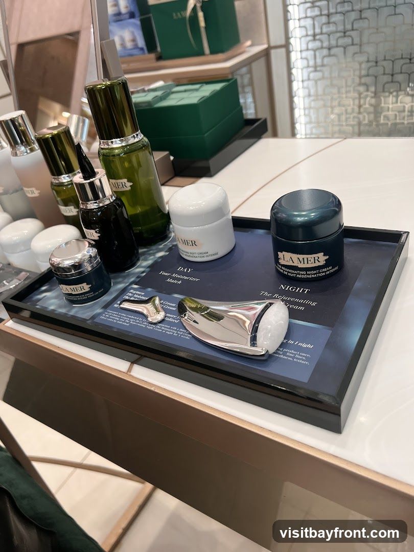 A display of La Mer skincare products, including creams and serums, arranged on a tray. The display features jars of "The Moisturizing Soft Cream" and "The Rejuvenating Night Cream," along with a silver facial massage tool. In the background, more La Mer products and branded boxes are visible on shelves.