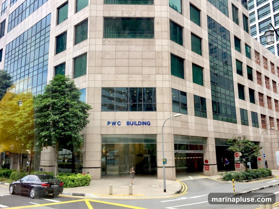 A view of the PWC Building's entrance and facade. The building has a modern design with large glass windows and beige stone cladding. The words "PWC BUILDING" are visible in blue lettering above the entrance. A black car is parked on the street in front of the building, and trees and greenery are planted along the sidewalk.