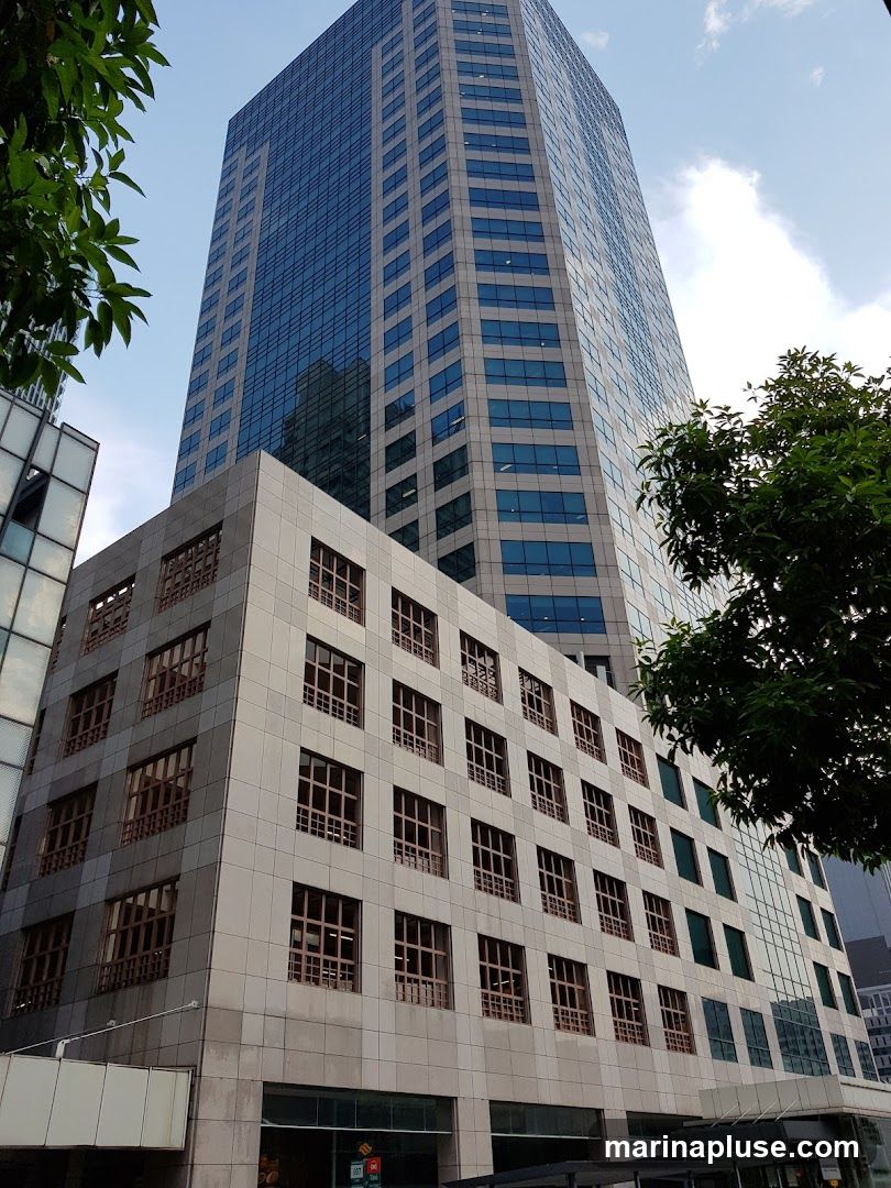 A low-angle shot of a modern skyscraper with a glass facade and a lower section clad in stone. Trees frame the building on the left and right. The sky is partly cloudy.
