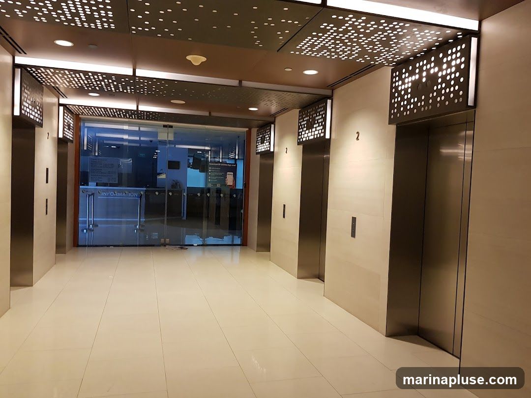 A modern elevator lobby with polished floors and a decorative ceiling. There are several elevators with illuminated panels above them. To the left, a glass-enclosed information desk is visible with signage that includes the word "INFORMATION".