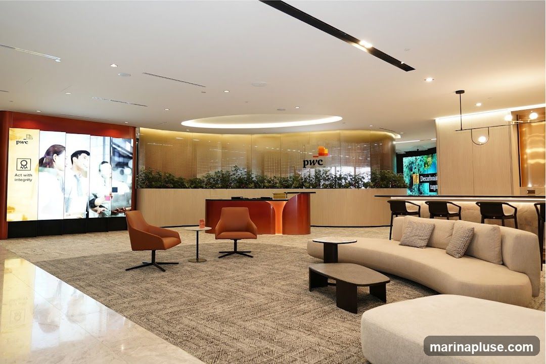 A modern and spacious reception area with a large digital display on the left showing the PwC logo and the text "Act with integrity." In the center, a curved reception desk with orange accents is visible, behind which is a wall with plants and the PwC logo. To the right, a bar area with stools and a curved sofa with cushions are arranged. The flooring is carpeted with a marble accent near the entrance.