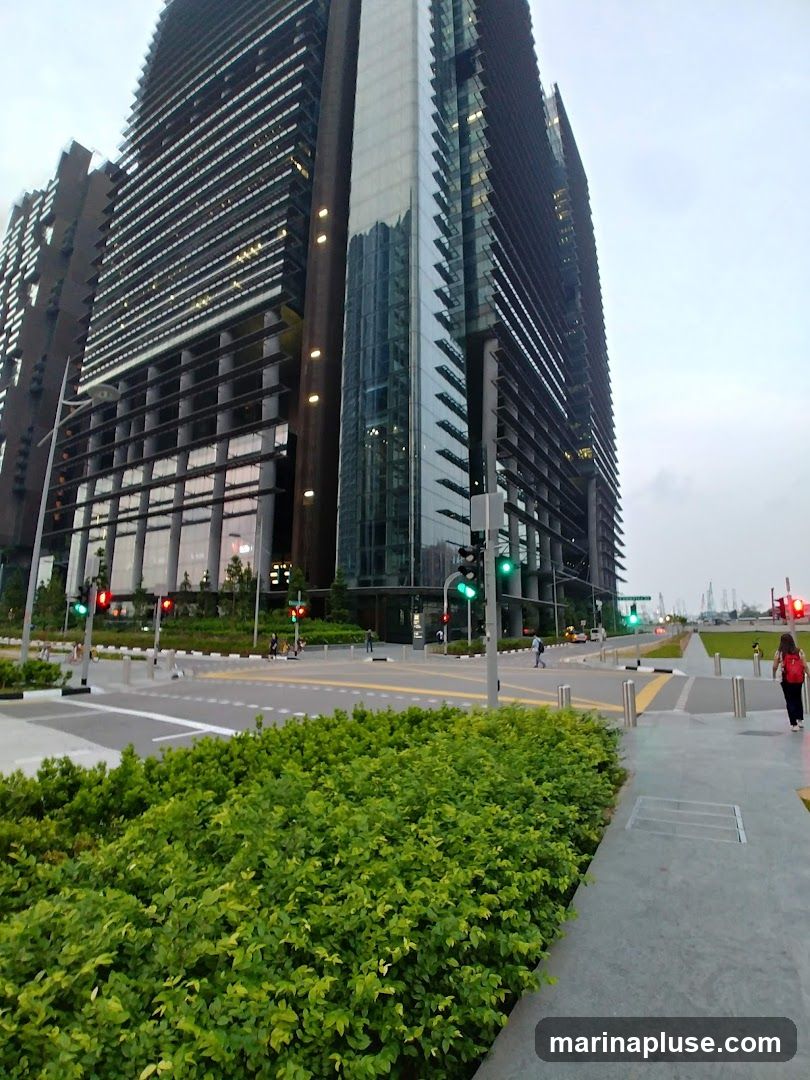 A wide-angle shot captures a modern, multi-story skyscraper with a distinctive facade featuring horizontal louvers and glass panels. The building is illuminated, with lights visible through the windows. In the foreground, lush green bushes line a paved walkway and a street intersection with traffic lights. Several people are visible walking on the sidewalks and crossing the street. The sky is overcast, suggesting it might be early morning or late afternoon.