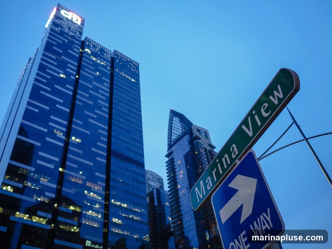 A low-angle shot of modern skyscrapers against a clear blue sky. The tallest building on the left features the 'citi' logo illuminated at its peak. Street signs for 'Marina View' and 'ONE WAY' are prominently displayed in the foreground, indicating a street intersection. The windows of the skyscrapers reflect the twilight sky and city lights.