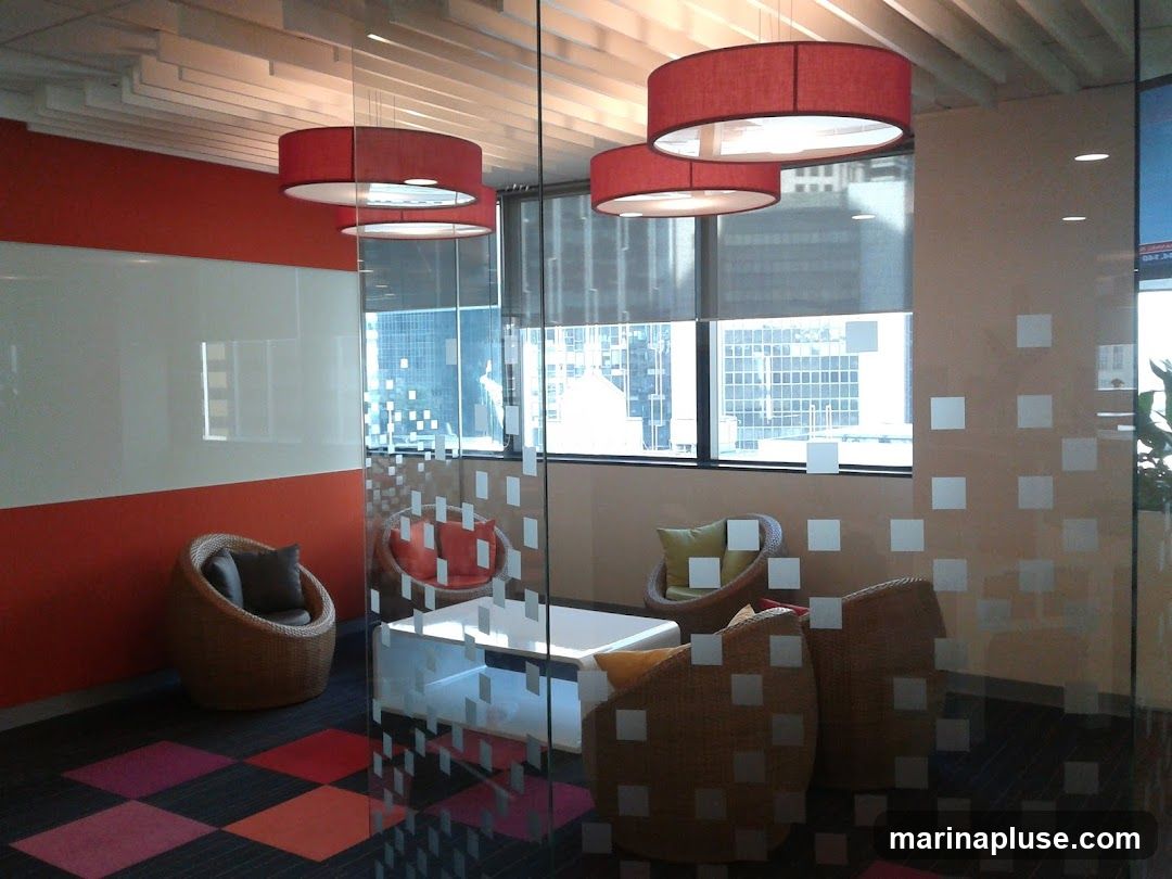 A contemporary meeting room with a glass partition, featuring woven chairs, a white coffee table, and large red pendant lights. The walls are painted orange and white, and the floor has a colorful checkered carpet. Large windows offer a view of city buildings.