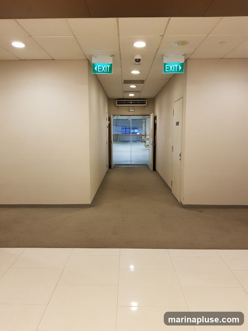 A view down a carpeted hallway with white walls. Two green illuminated exit signs are visible hanging from the ceiling, one on the left pointing left and one on the right pointing right. A security camera is mounted on the ceiling between the signs. At the end of the hallway, glass doors lead into a brightly lit room with blue accents. A white door is visible on the right side of the hallway. The floor in the foreground is tiled with white squares.