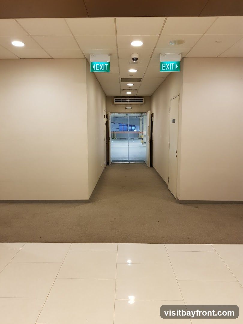 A view down a carpeted hallway with white walls. Two green illuminated exit signs are visible hanging from the ceiling, one on the left pointing left and one on the right pointing right. A security camera is mounted on the ceiling between the signs. At the end of the hallway, glass doors lead into a brightly lit room with blue accents. A white door is visible on the right side of the hallway. The floor in the foreground is tiled with white squares.