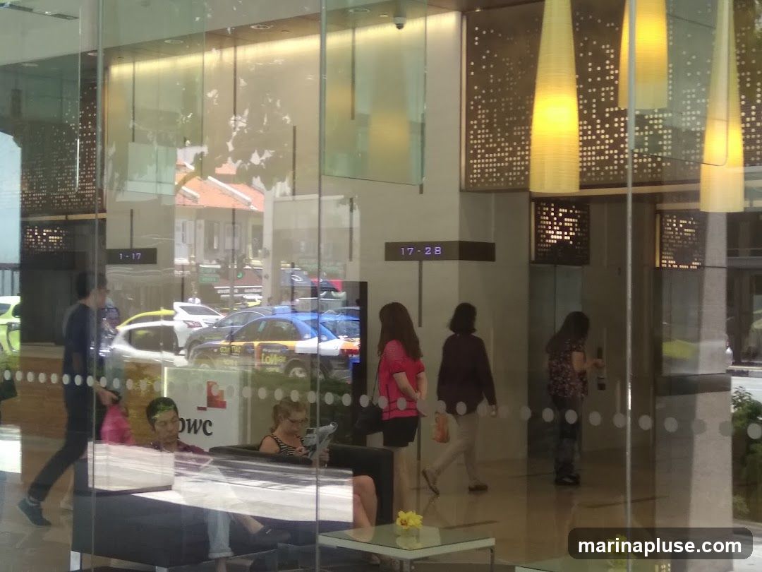 A view through a glass facade into a modern lobby. People are seen walking and sitting. Reflections of the street, including cars and buildings, are visible on the glass. Large, decorative pendant lights hang from the ceiling. Digital displays show times. A logo with the letters "owc" is visible on a panel inside.