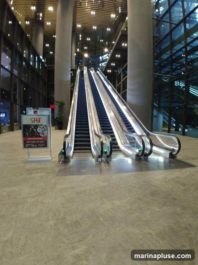 A wide shot of multiple escalators ascending in a spacious, modern building. The escalators are illuminated and have clear glass railings. Large pillars support the ceiling, which is lined with lights. The floor is a polished concrete or stone. To the left, a sign with the logo "SRT" is visible, displaying images of food and text. The overall impression is one of a clean, contemporary commercial or public space.
